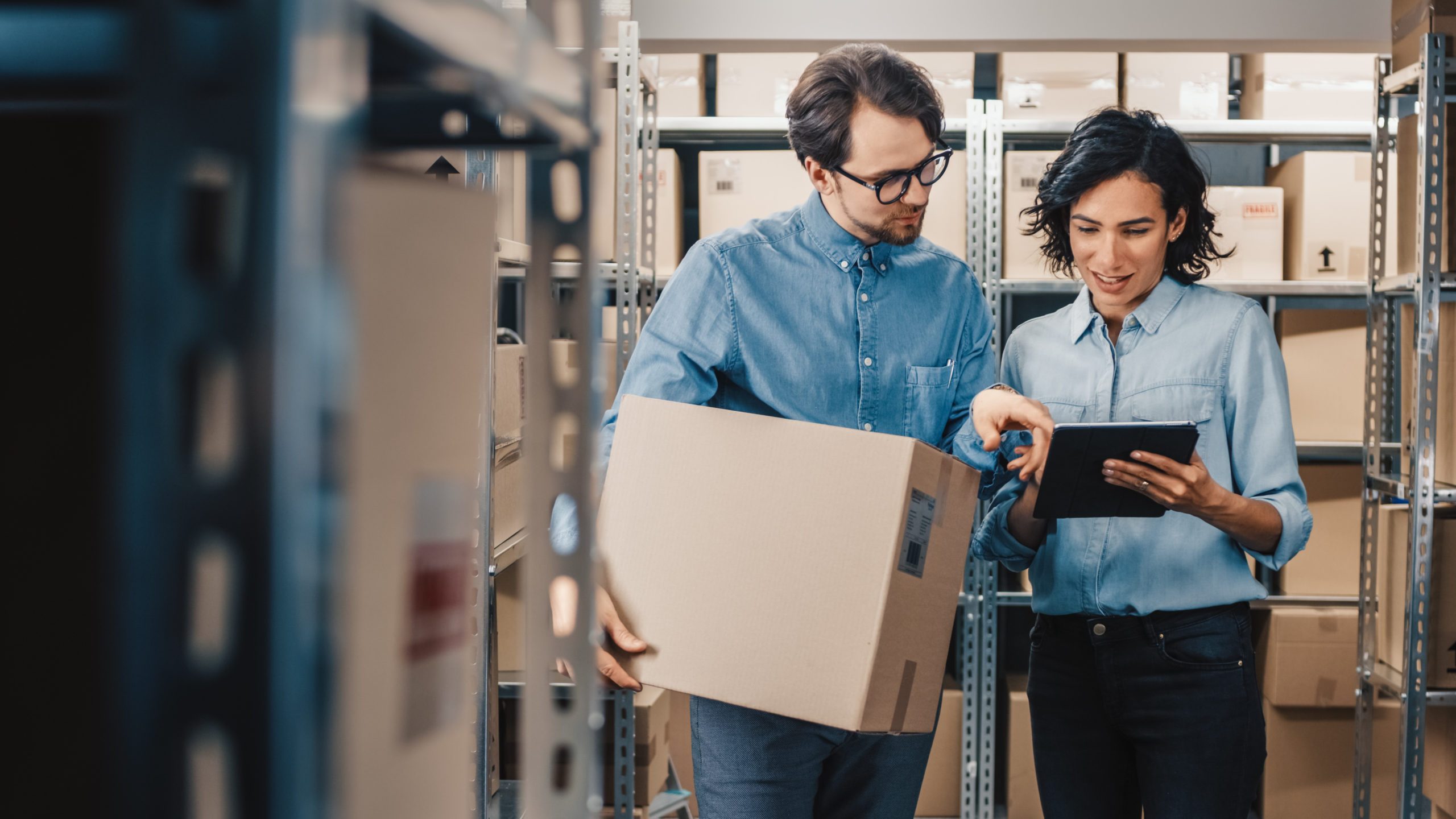 Female Inventory Manager Shows Digital Tablet Information to a Worker Holding Cardboard Box, They Talk and Do Work. In the Background Stock of Parcels with Products Ready for Shipment. 领航国际官方网站图片